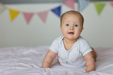 Wall Mural - Caucasian blonde baby six months old lying on bed at home. Kid wearing cute gray clothing. Bedroom decorated with flags.