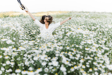 Wall Mural - portrait of a beautiful young Caucasian woman on a chamomile field in a picturesque valley. a young lady in a white dress and a straw hat jumps out of a bush.