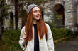 © alfa27 - Close-up Portrait Of Dreamy Woman With Natural Red Hair On Travel Trip, Outdoors, Posing At Camera, Smiling, in Coat And Hat. Old Building In The Background