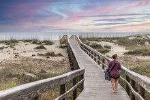 A Woman Walking Alone On The Wooden Walkway Path Out To The Ocean Ar Tybee Island At Sunset.