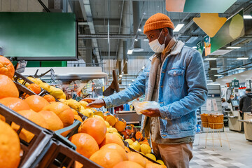 Wall Mural - African-American man customer in warm jacket with disposable mask takes fresh lemon fruits from crate in supermarket side view