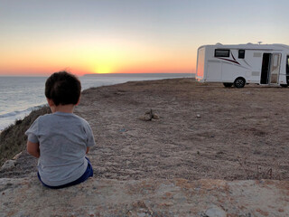 Child on his back sitting looking at the sunset by the sea near a caravan.