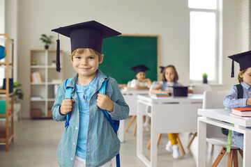 Portrait of happy school child. Successful elementary school graduate in academic grad hat standing in classroom. Cute little boy in mortarboard looking at camera and smiling