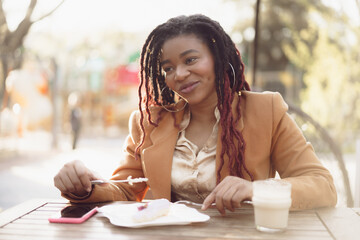 Smiling african american woman drinking coffee and eating dessert in outdoor cafe