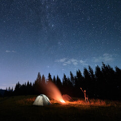 Wall Mural - man hiker near campfire and pointing finger at night sky with stars. picturesque view of blue starry