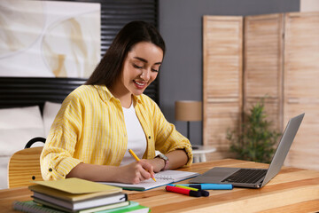 Wall Mural - Young woman writing down notes during webinar at table in room