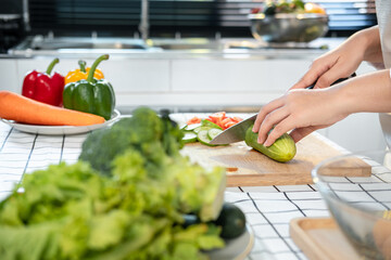 Wall Mural - Asian housewife wearing apron and using knife to slice cucumber and tomato on chopping board