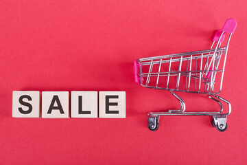 Mini supermarket cart and wood cubes with text SALE on red background. Online shopping, business concept. Top view, flat lay with copy space.