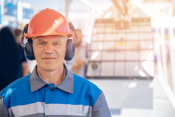 Industrial worker engineer man in protective helmet and headphones in uniform on background of factory sunlight