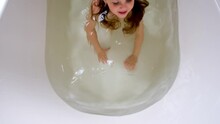 Funny Little Girl With Long Wet Hair And Blue Eyes Jumps In Water In Large White Bath Under Bright Electric Light Close Upper View