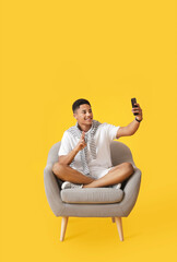 Canvas Print - Young African-American guy with phone sitting in armchair on color background