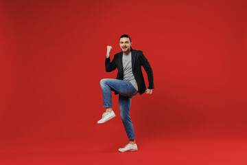 Wall Mural - Full length young businessman latin man 20s with long hair ponytail wearing black striped jacket grey shirt look camera do winner gesture with raised up leg isolated on red background studio portrait