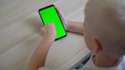 Caucasian boy in a gray T-shirt sits at a table and holds a phone with a green screen in his hands. The child presses his finger on the phone screen while playing games or using an application.