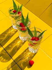 Wall Mural - Refreshing summer drinks in crystal glasses with green leaves raspberry yellow tile background shadows. Sweet cold natural cocktail hot summer day. Soda water fruits berries mint tonic. Party vacation