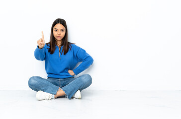 Young mixed race woman sitting on the floor isolated on white background counting one with serious expression