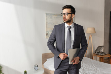 Wall Mural - Young businessman in suit and eyeglasses holding laptop in bedroom