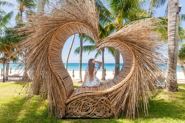 Back View of Tourist Woman Sitting in Photo Spot like Straw Heart with Tropical Palm Trees and Blue Sea with White Sandy Beach on Background. Nature and travel concept