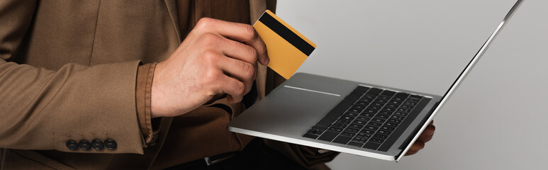 Wall Mural - Cropped view of man in formal wear holding credit card and laptop isolated on grey, banner