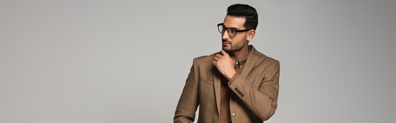 Poster - Arabian businessman in eyeglasses and brown jacket looking away isolated on grey, banner