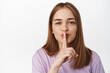 © Cookie Studio - Hush. Close up portrait of blond girl shushing, smiling and asking to keep quiet, has secret, shhh gesture, press finger to lips, has surprise, standing against white background