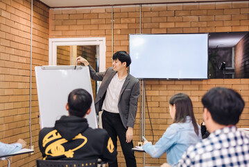 business cooperation : Young asian male coach or speaker make flip chart presentation to diverse businesspeople at meeting in office. Male tutor or trainer present project to diverse colleagues.