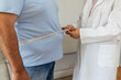 © Jelena Stanojkovic - Cropped shot of a female doctor taking obese patient's body fat measurements. Dietitian measuring abdominal circumference during checkup. Young doctor taking measurement from depressed overweight man.
