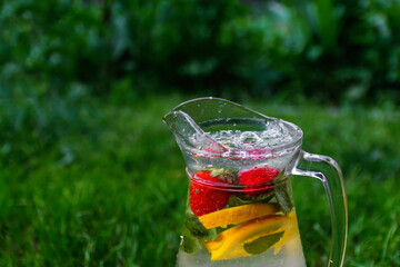 Defocus glass jug of lemonade with slice lemon and mint on natural green background with water drops. Pitcher of fresh cold ice summer cocktail or punch. Drops, wet, bubbles. Copy space. Out of focus