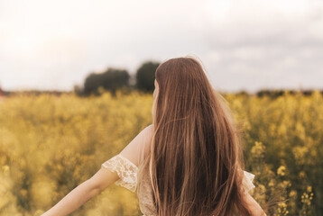 A young beautiful girl with long hair walks in a rapeseed field, looks into the distance. rear view.