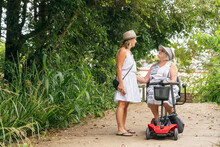 An Elderly Woman On A Disability Scooter With Her Daughter Walking Down A Path In A Park