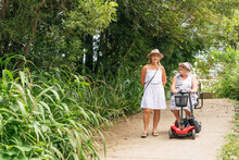 An Elderly Woman On A Disability Scooter With Her Daughter Walking Down A Path In A Park
