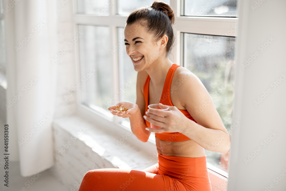 Happy woman takes supplements or vitamins in the form of capsules after training in the gym. Concept of additional nutrition during training - obrazy, fototapety, plakaty 