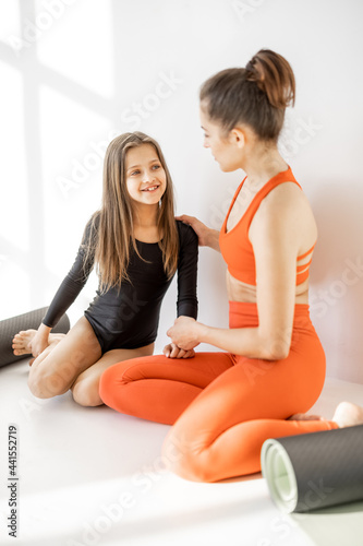 Sports mom and little daughter sitting together with yoga mats and have a close conversation during sports indoors. Close relationships with mother and sports in childhood