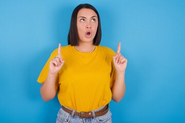 young beautiful brunette girl with short hair standing against blue background amazed and surprised looking up and pointing with fingers and raised arms.