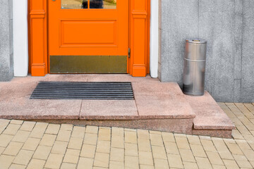 granite threshold with foot mat near orange wooden front door with iron trash can near building facade with gray cladding and yellow stone tile pavement close-up, nobody.