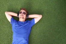 Close Up Portrait Of Smiling Man Lying On Grass  While Listening To Music An Wearing Sunglasses.Happy Man Lying With Both Hands Resting Behind His Neck On The Grass