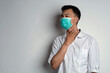 Portrait of young asian man wearing health mask having sore throat and touching his neck using his right hand, against white background. Man having sorethroat. Novel Coronavirus (COVID-19)