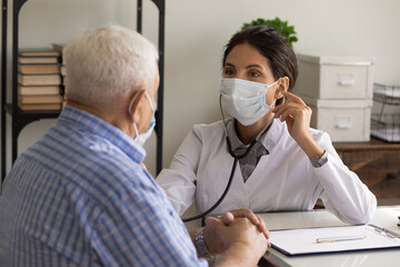 Wall Mural - Young female doctor in facemask listen to old male patient heart chest with stethoscope at clinic meeting. Woman GP checkup examine mature man client with phonendoscope. Elderly healthcare concept.