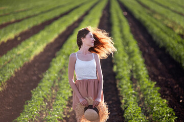 Young woman in white T-shirt, powdery shorts runs along green plowed field. Long hair develops at sunset, the girl has straw hat. Promote agricultural perspective, the texture of black soil ground