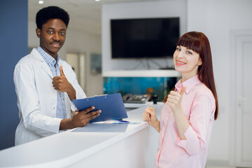 Wall Mural - Medical male afro-american worker consults beautiful young woman patient in modern medical clinic. Patient and doctor standing near the reception and showing thumbs up