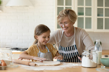 Wall Mural - Happy diverse generations female family having fun, involved in preparing homemade pastry together in modern kitchen. Joyful old granny sharing culinary skills with cute little granddaughter.