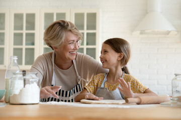 Wall Mural - Happy mature older grandmother having fun with adorable small kid granddaughter, enjoying cooking together in modern kitchen. Joyful different generations family involved in culinary activity.