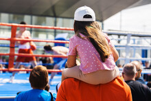 A Little Girl Sits On Her Father's Neck And Watches A Boxing Competition. Street Fights In The Ring. Unrecognizable People