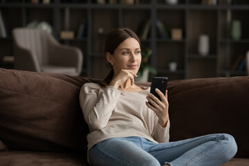 Dreamy young beautiful woman looking in distance, thinking of pleasant mobile message. Happy relaxed caucasian lady holding smart phone in hands, getting pleasant news, visualizing future at home.