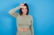 Funny Young beautiful sporty girl standing against blue background makes loser gesture mocking at someone sticks out tongue making grimace face.