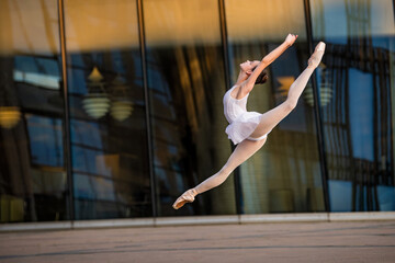 Wall Mural - young ballerina in a white leotard is dancing on pointe shoes against the background of a cityscape, frozen in jump