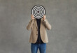 Unknown businessman hid his face behind a darts board while standing on a gray background. Concept of setting business goals, finding the target audience, achieving goals and success