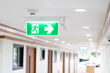 A Arrow Light Box Sign Of EMERGENCY FIRE EXIT Is Hung On The Ceiling In Hospital Walkway, Idea For Event Fire Or Evacuation Drills.