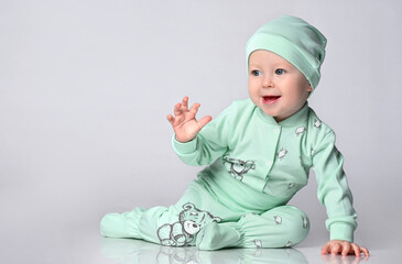 joyful baby toddler The toddler sits in a cotton jumpsuit with a print of patches and a matching cap, raised his hand and waves hello or goodbye.