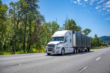 Bonnet big rig semi truck in white color and with chrome grille guard transporting cargo in tented dry van semi trailer driving on the wide multiline road