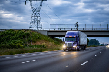 Bright blue big rig semi truck transporting cargo in refrigerated semi trailer running under the bridge on the wide highway road at the night time